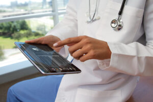 * Photo by NEC Corporation of America with Creative Commons license. Photo shows a cropped image of a person in a lab coat and wearing a stethoscope. They are holding a computer tablet and pointing at the screen.