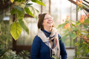 Giskin Day, with medium length brown hair, scarf and glasses, laughing in bright light. Botanical plants in the fore and background.