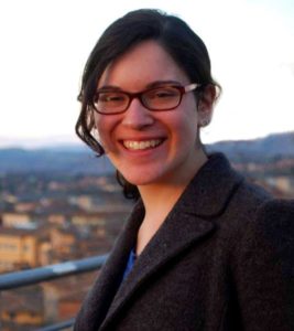 Ranana Dine, a brunette with glasses in a tweed jacket, smiling with a cityscape in the background.