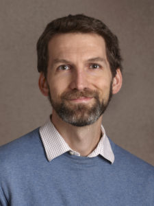 A headshot of the author, light brown hair and a trim beard. He wears a blue sweater and collared shirt, and smiles slightly.