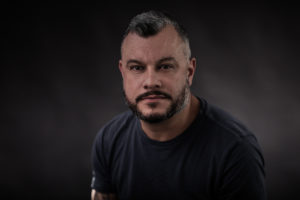 Colot photo from shoulder of Steve Wise. He wears a short-croppsed mustache and bead, close cut hair and black shirt. Facing the viewer.