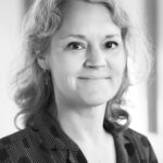 A black and white photo of Anna, who has shoulder-length blond (curly) hair. She wears a patterned blouse and smiles, turned three-quarters toward the camera.