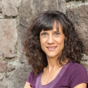 Image description for the alt text: Headshot of a white woman with olive complexion and curly brown hair. She stands against a rough stone wall and smiles slightly