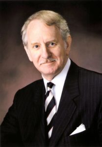 A head shot of Dr. Cooper. David has gray hair and trim mustache. He wears a suit and black and white striped tie against a muted background and smiles slightly.
