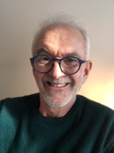 Headshot of Hugh, smiling in sunlight. He has a short gray beard and glasses, and wears a green shirt. 