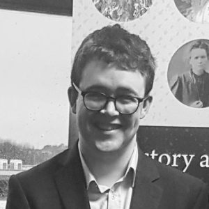 A black and white photo of David, smiling in sunshine. He wears glasses, short-cropped hair, and a collared shirt.