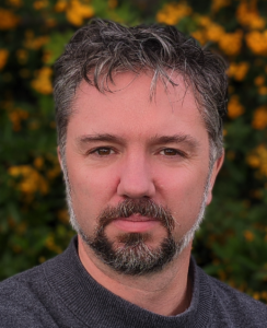 A head shot of James with fall color int he background. He wears a short beard and salt-and-pepper close-cropped hair. He smiles slightly., 