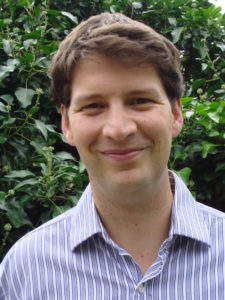 Joshua wears a striped blue shirt and stands before a backdrop of lush greenery. He smiles slightly.