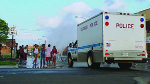 A police vehicle in front of a fire hydrant soaking the street; a small group of people gather to the left