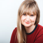 Head shot of Vanessa; she wears a red shirt with long blonde hair over one shoulder against a white background. She smiles slightly.