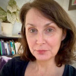 A headshot with bookshelf in background. She has brown shoulder-length hair and smiles slightly