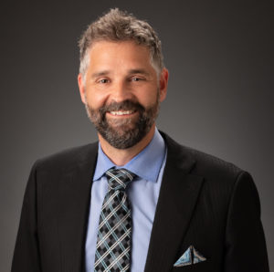 A headshot of Josh Mugele against a dark gray background. He wears a sportcoat and blue shirt with tie, has a beard and salt-and-pepper hair. He smiles at the camera.