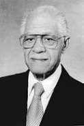Black and White Head Shot of an older Dr. Watts. He wears a suit and glasses and siles slightly.
