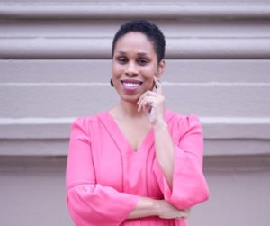 Headshot of Oni Blackstock; she weats a pink blouse and raises one hand to the left of her face. She is smiling in front of a gray background.